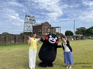 三池炭鉱万田坑の前でインタビューの方と笑顔で写真を撮るくまモンとアナウンサー
